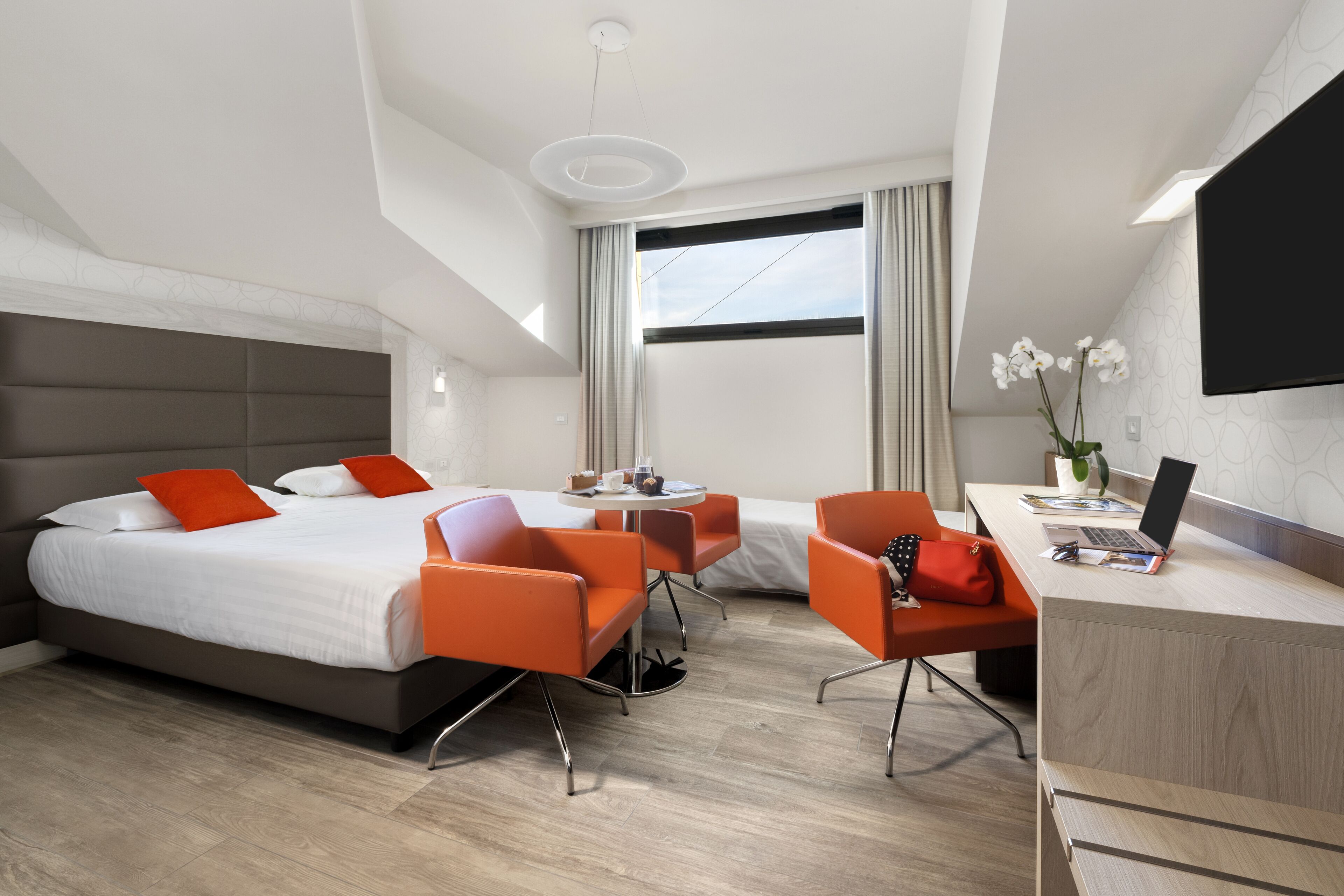 Ein modernes Hotelzimmer mit einem großen Bett, zwei orangefarbenen Sesseln, einem Schreibtisch mit einem Laptop und einem Fernseher.
