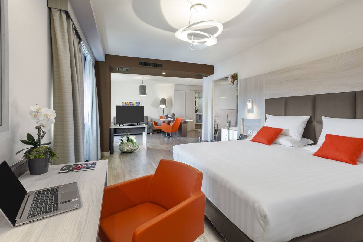 Habitación de hotel moderna con una cama grande, un escritorio con una computadora portátil, una silla naranja y vista al comedor.
