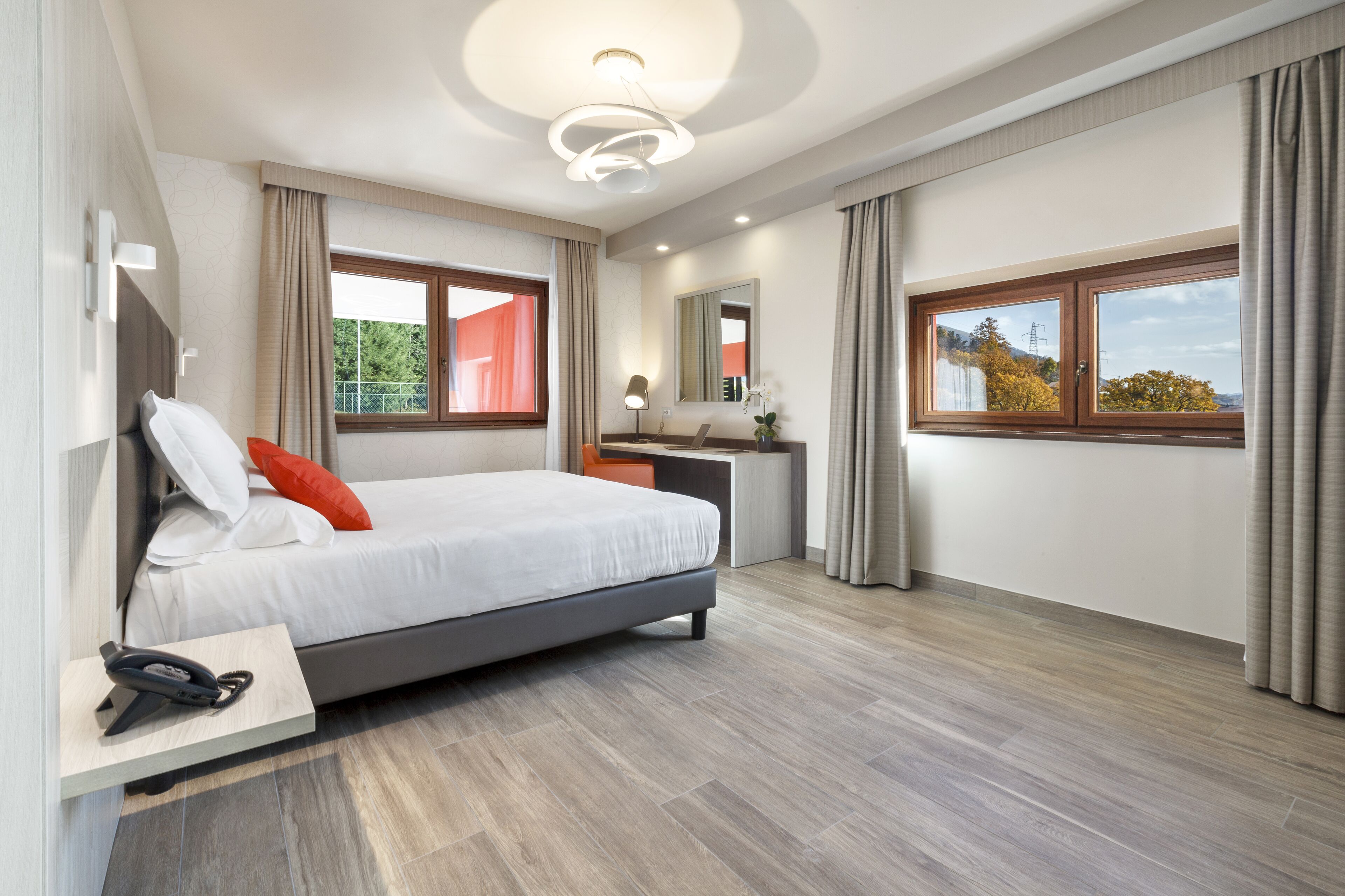 A modern hotel room with a bed, bedside table, lamp, and a view of the outdoors through the windows.