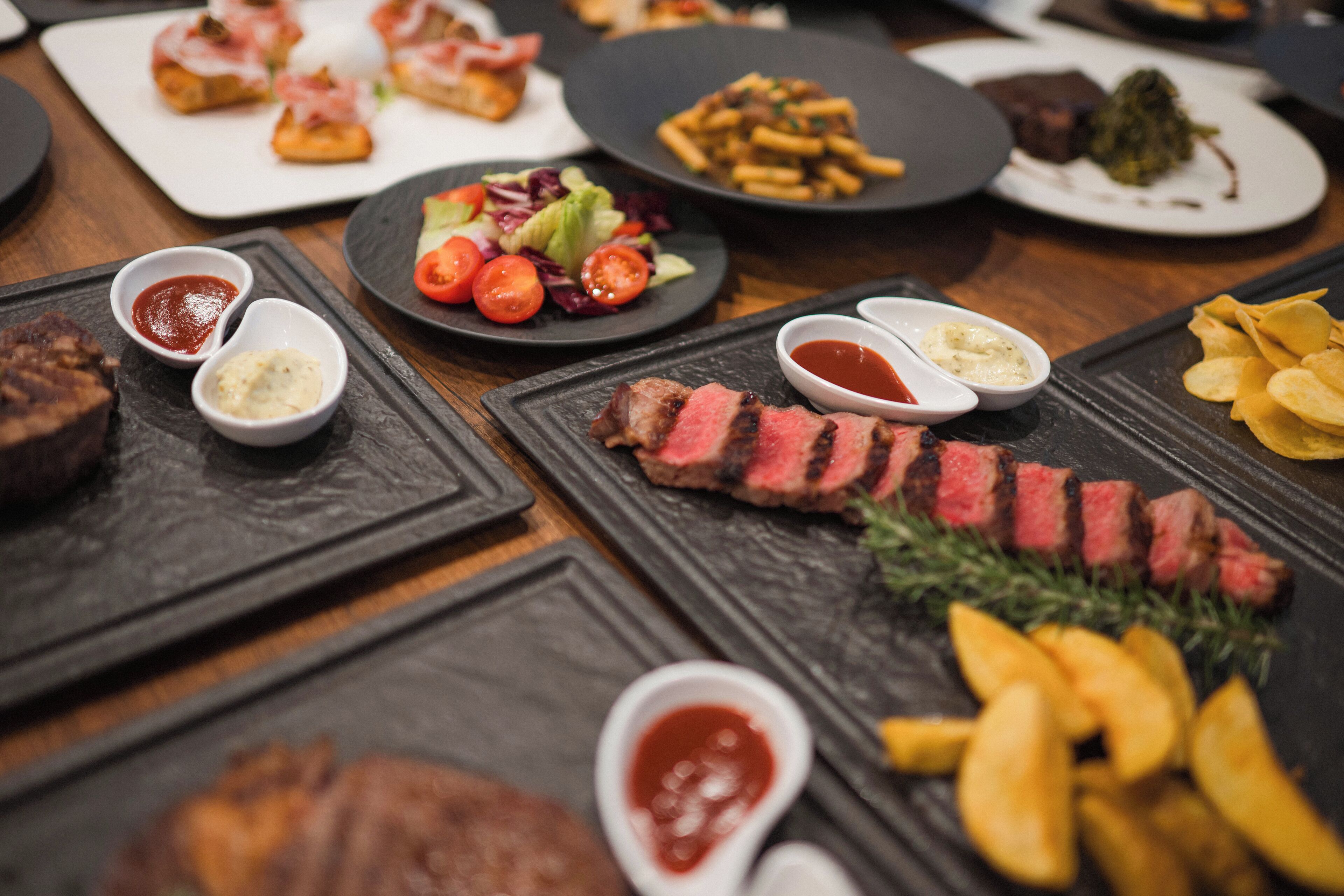Diversos pratos, incluindo bife, batatas fritas e saladas, sobre uma mesa de madeira.