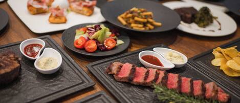Diversos pratos, incluindo bife, batatas fritas e saladas, sobre uma mesa de madeira.