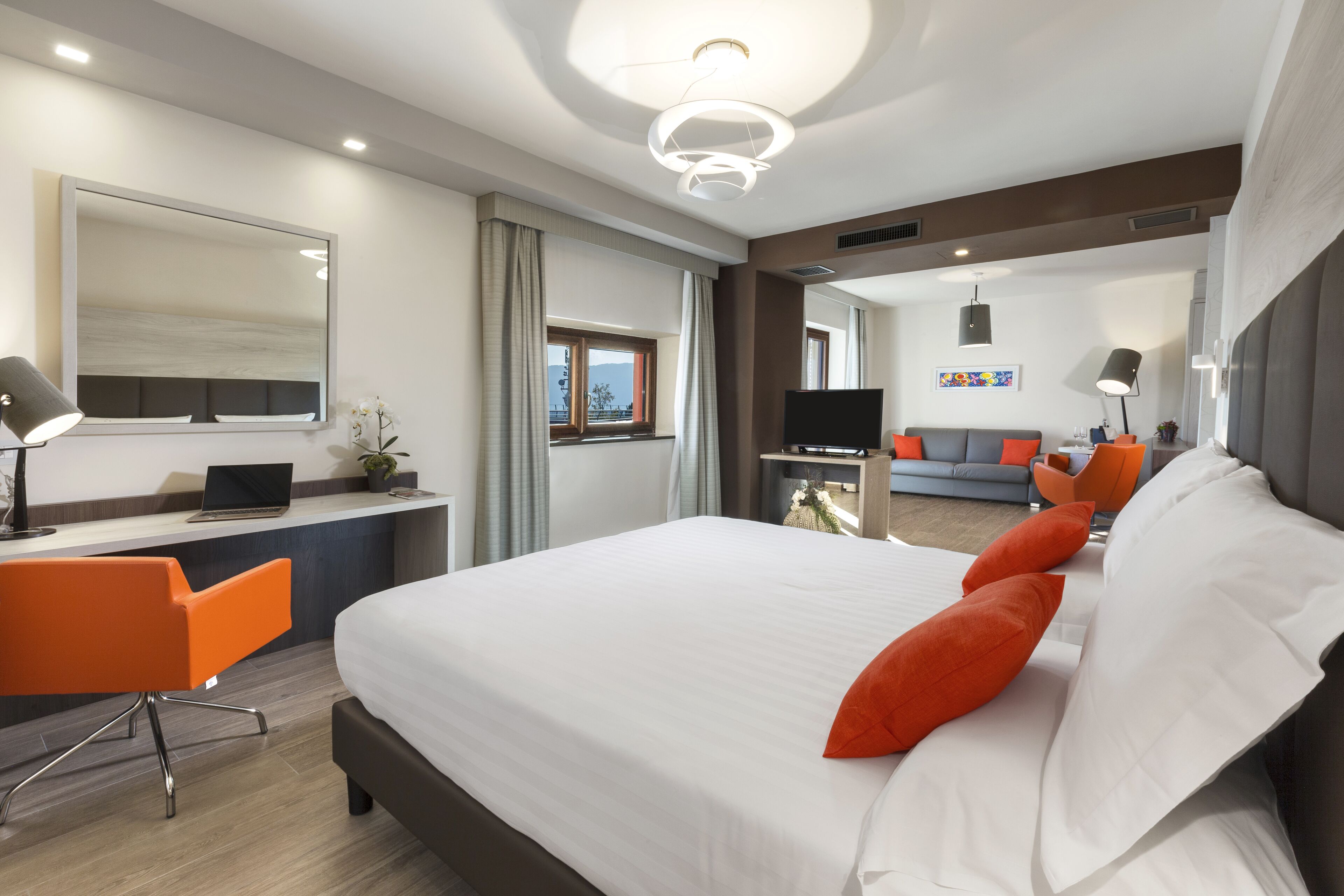 A modern hotel room with a large bed, an orange chair, a desk with a laptop, a TV, and a sofa in the background.