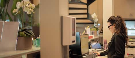 A woman working at a desk with a computer, surrounded by office supplies and potted plants.