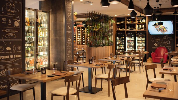 A restaurant with wooden tables and chairs, a wine cooler, and a television screen displaying food. - BHotel (Lagonegro)