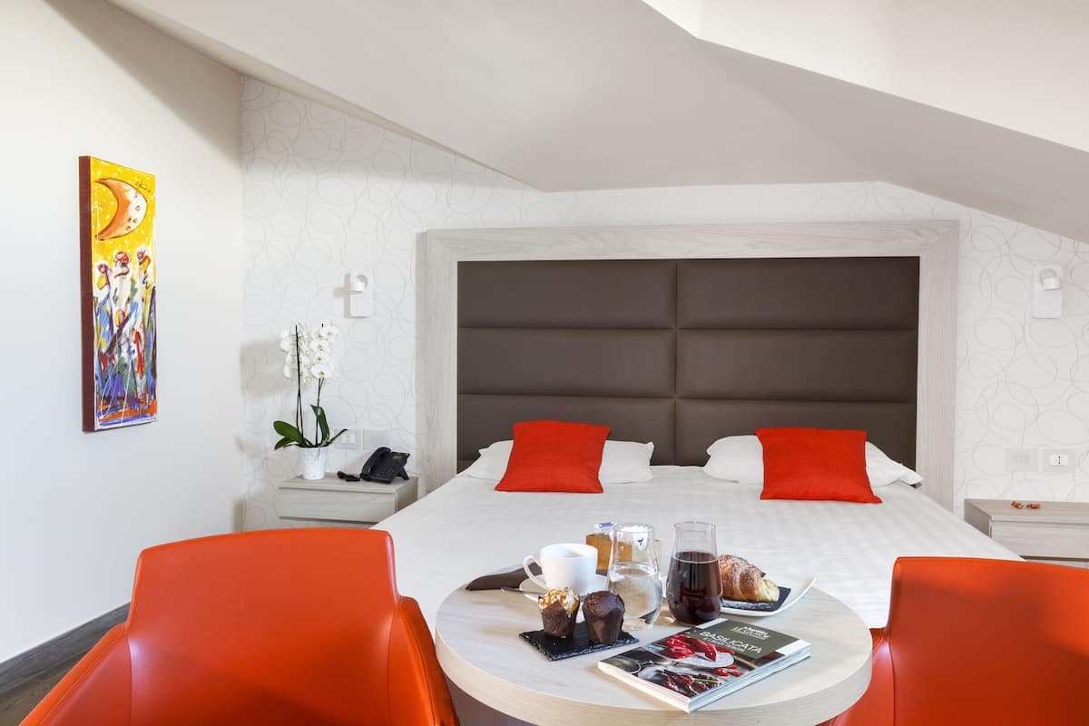 Una habitación de hotel moderna con una cama grande, almohadas rojas, una mesa de comedor con sillas naranjas y una mesita auxiliar con un teléfono y flores.