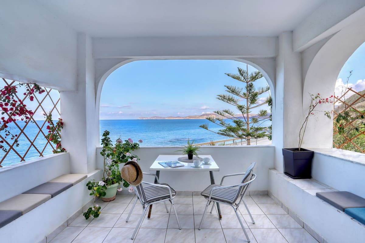 Un balcón con mesa y sillas, vista al mar y plantas en macetas.