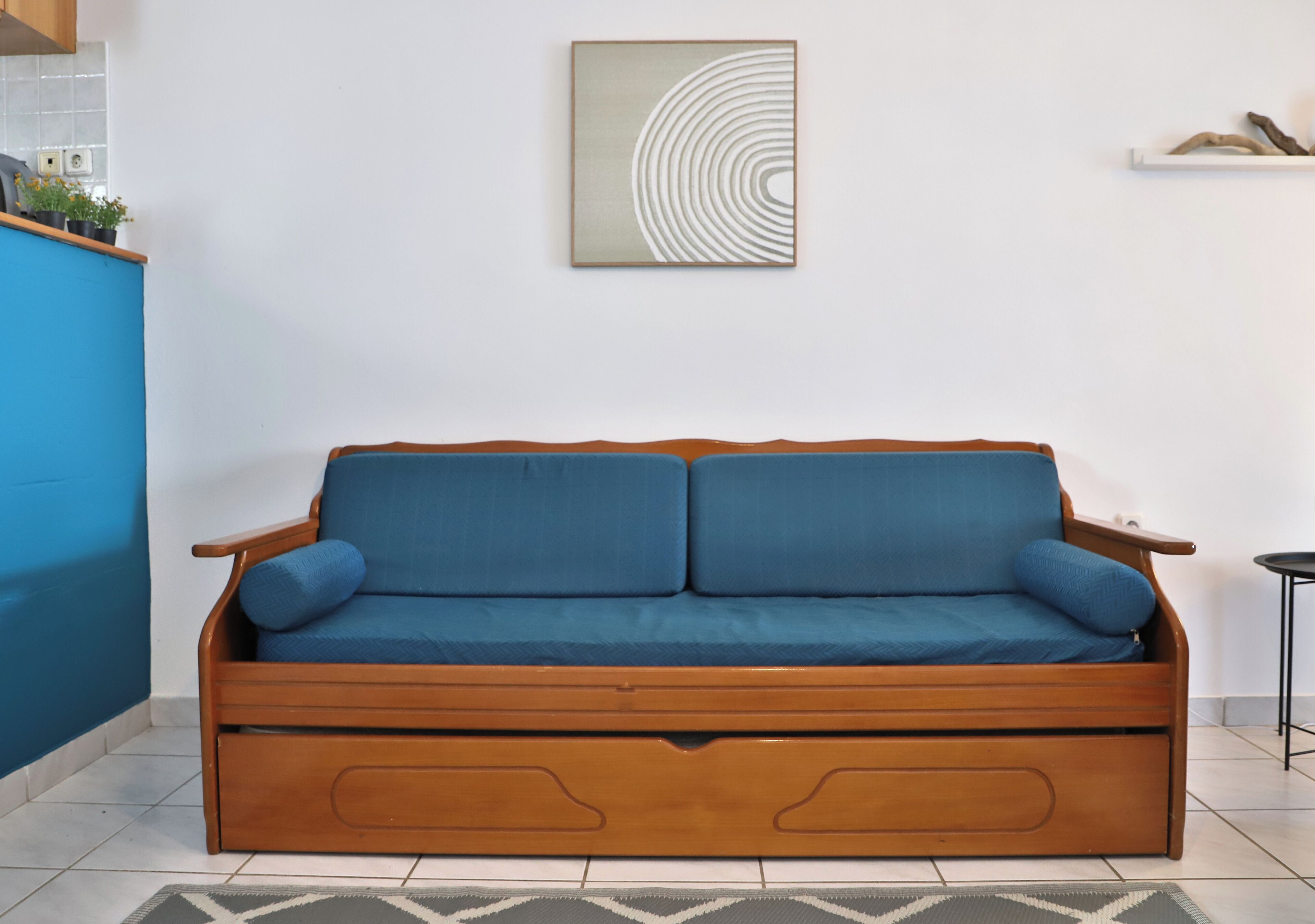 Een blauwe bank met houten armleuningen en lades, een witte muur met een cirkelvormig kunstwerk, en een klein plankje met decoratieve objecten.