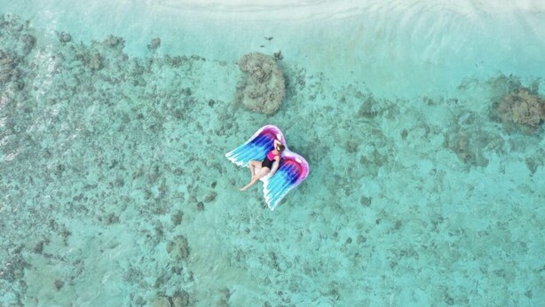 Una persona con una cola y alas coloridas sentada sobre una roca en una masa de agua azul cristalina.