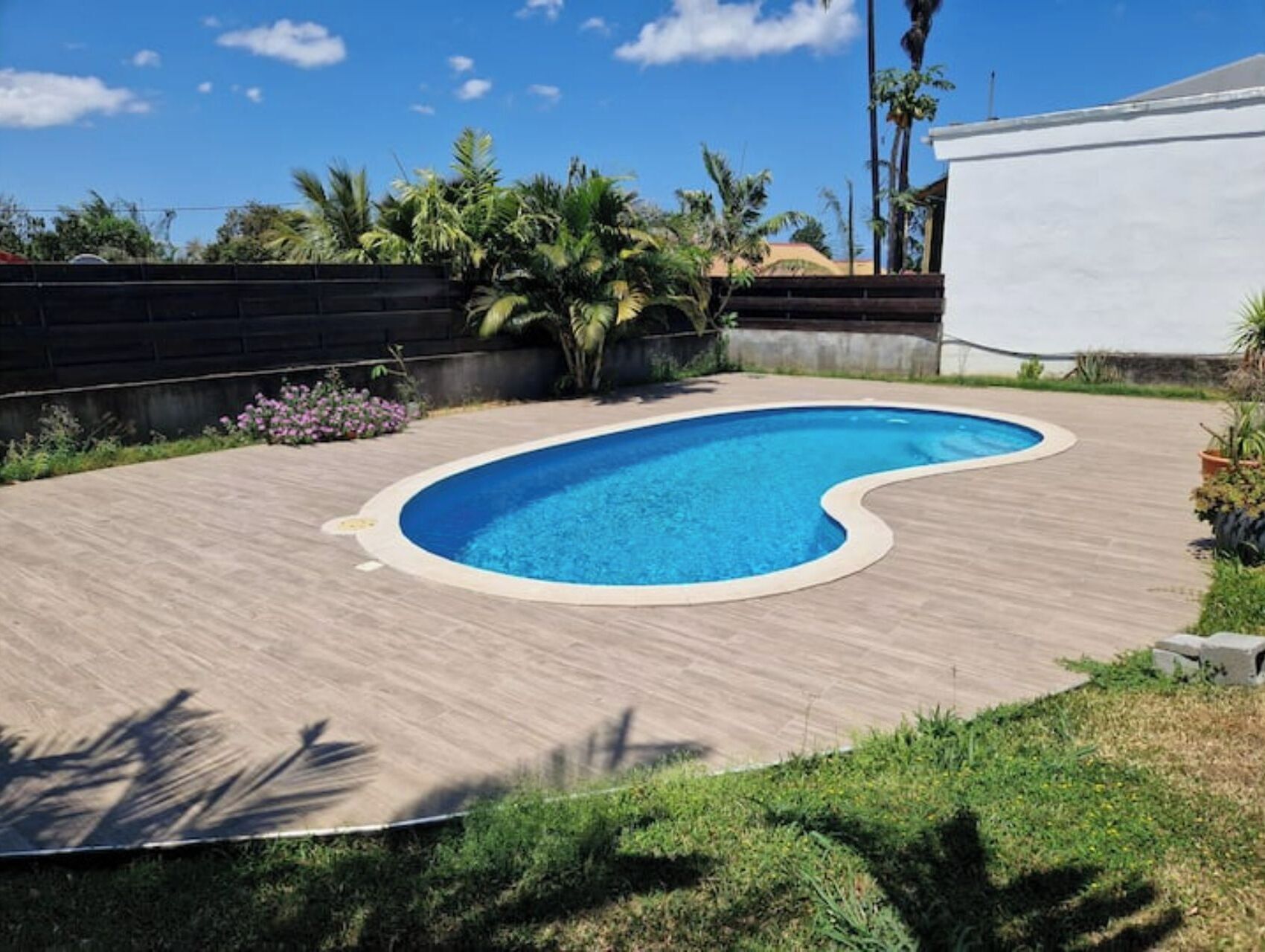 Une piscine dotée d’une terrasse en bois, entourée de verdure et bordée d’un bâtiment blanc.