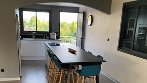 A modern kitchen with a central island, a dining area with teal chairs, and large windows offering a view of greenery. - Villa with swimming pool in the center of st louis river (Saint-Louis)
