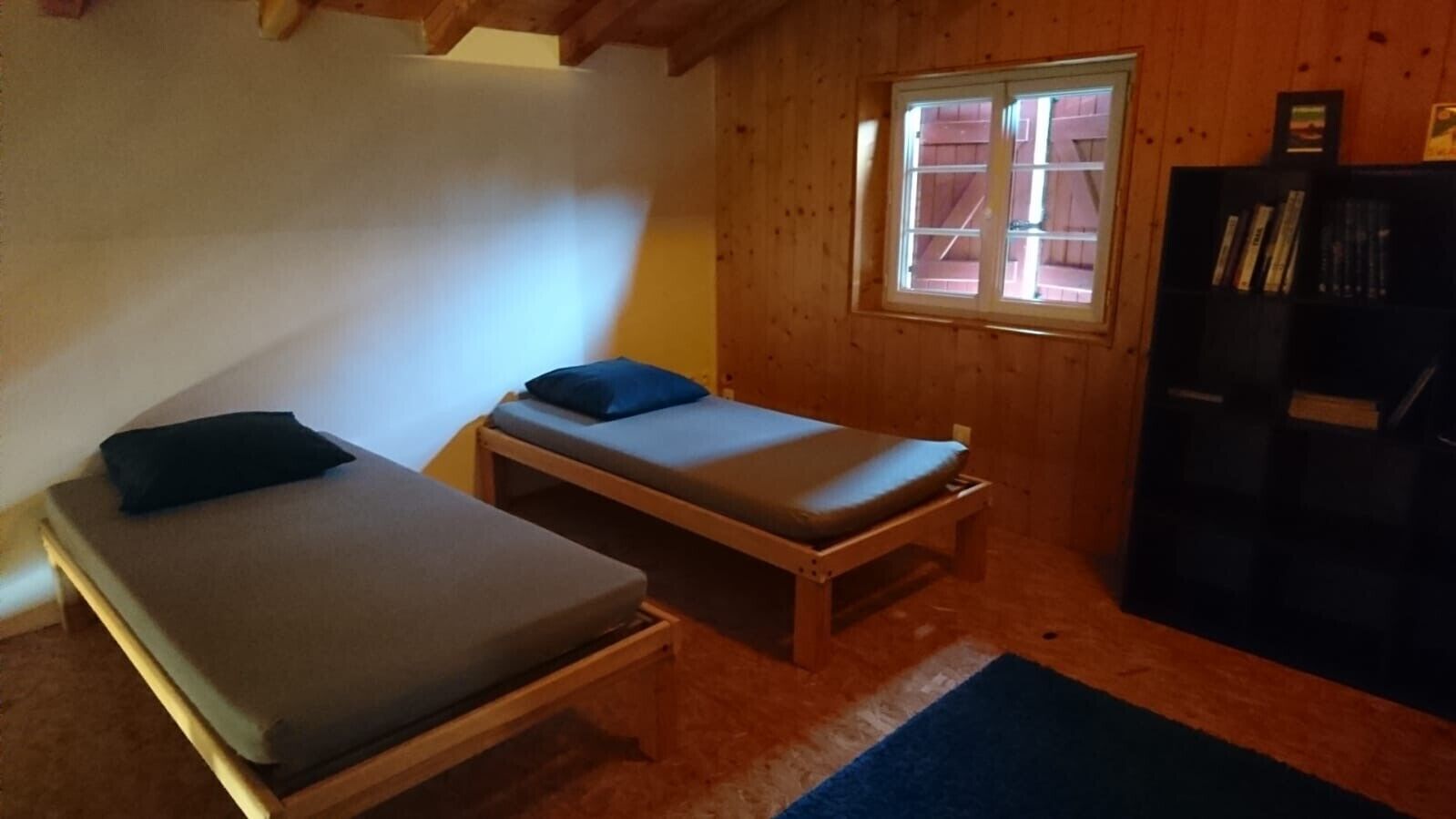 A small, cozy room with two wooden beds, a window, and a bookshelf.