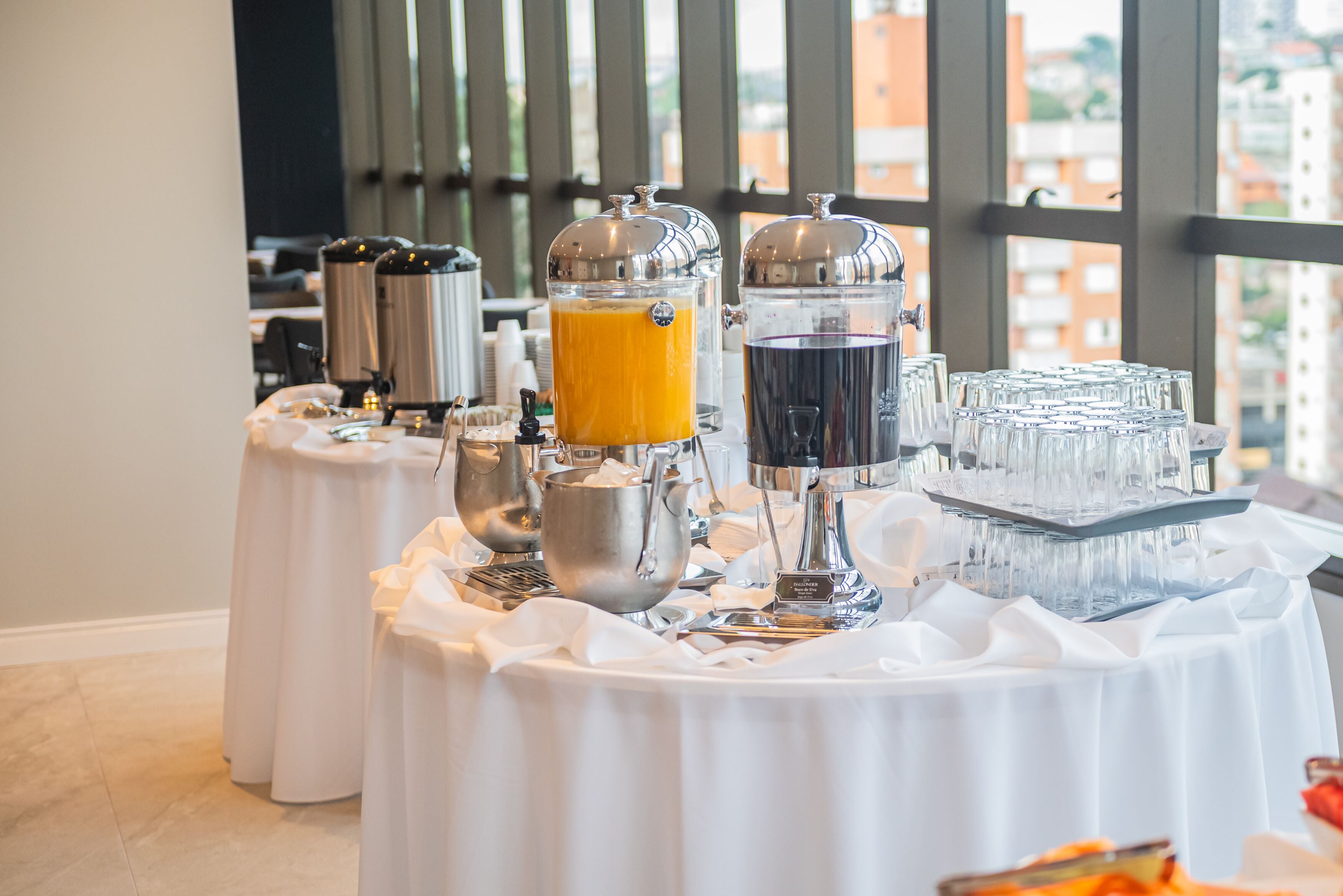 Disposição de buffet de café da manhã com diversos dispensadores de alimentos e bebidas, incluindo uma máquina de café, um dispensador de suco e um dispensador de chá, dispostos sobre uma mesa com toalha branca.