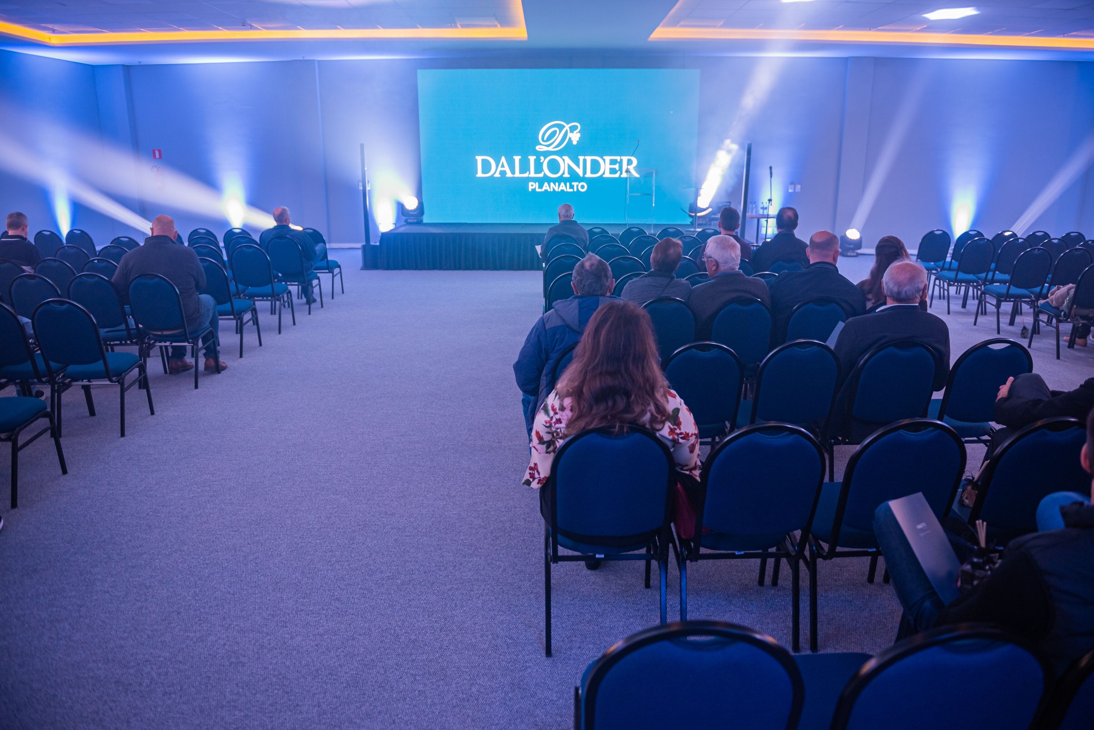 Un salón de conferencias con filas de sillas mirando hacia un escenario con una pantalla grande que muestra el texto "Dall'onder Planalto".