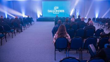 Uma sala de conferências com fileiras de cadeiras voltadas para um palco com uma tela grande exibindo o texto 'Dall'onder Planalto'.