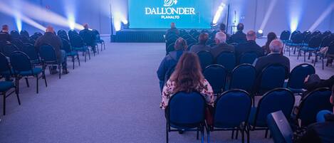 Un salón de conferencias con filas de sillas mirando hacia un escenario con una pantalla grande que muestra el texto "Dall'onder Planalto".