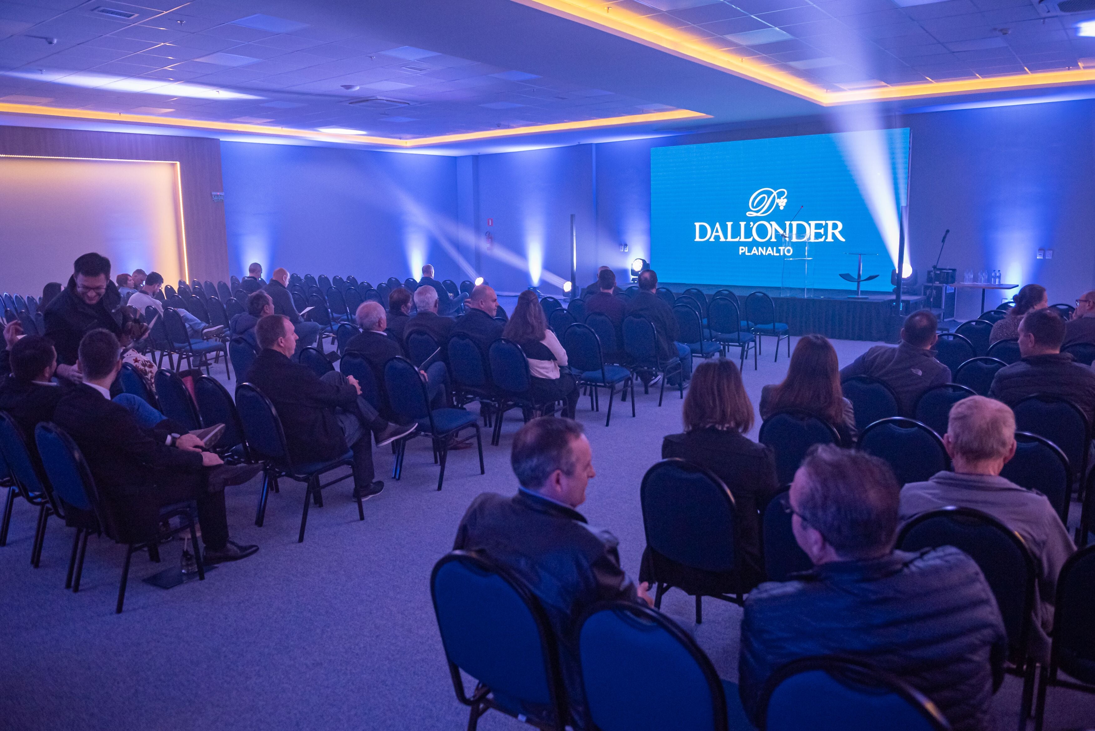 A conference room with rows of chairs facing a stage with a large screen displaying "DALLONDER PLANALTO".