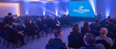 A conference room with rows of chairs facing a stage with a large screen displaying "DALLONDER PLANALTO".
