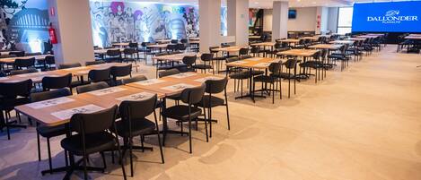 A spacious dining area with multiple tables and chairs, a large mural on the wall, and a screen displaying the hotel name.