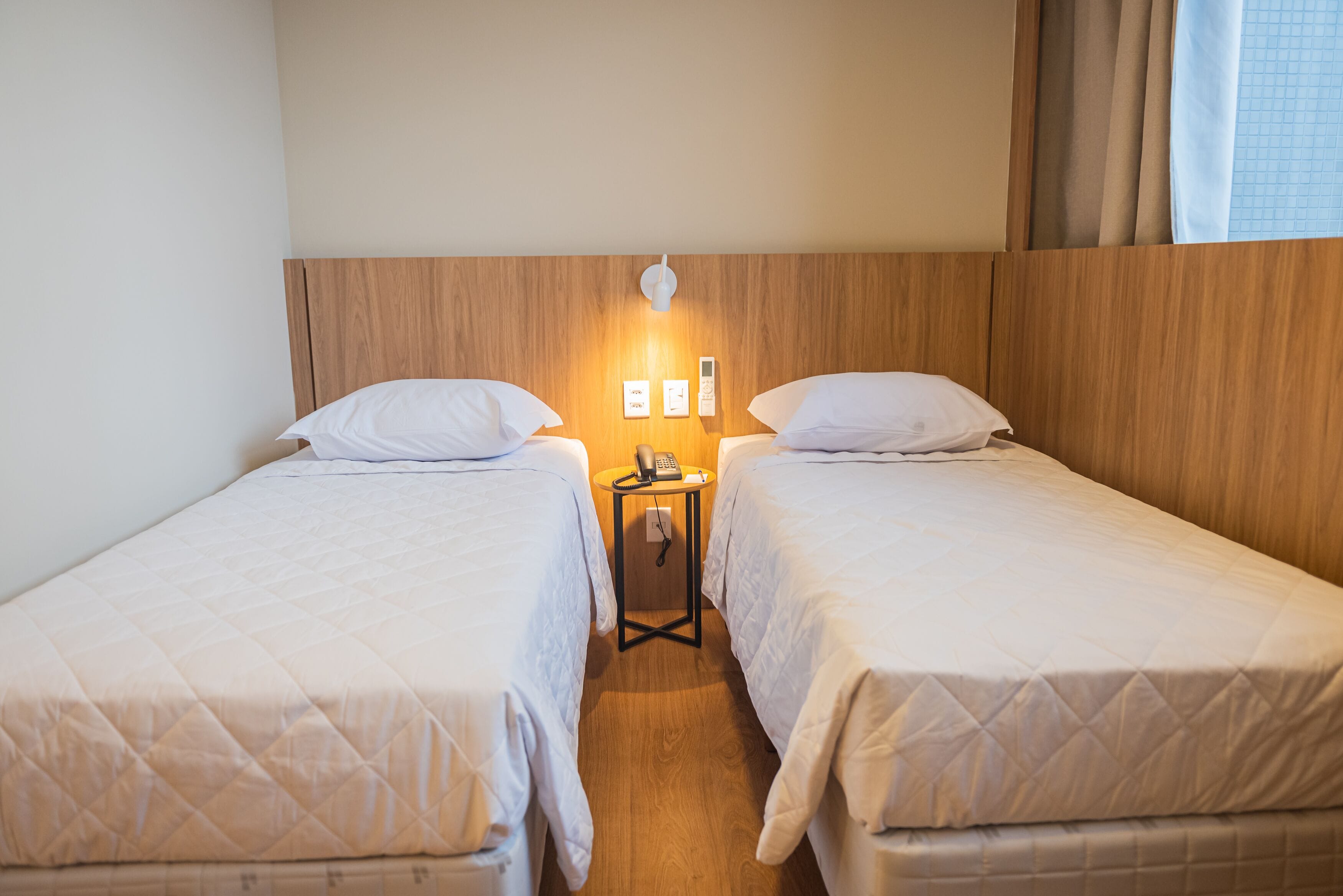 A hotel room with two single beds, a bedside table with a phone, and a wall-mounted light.