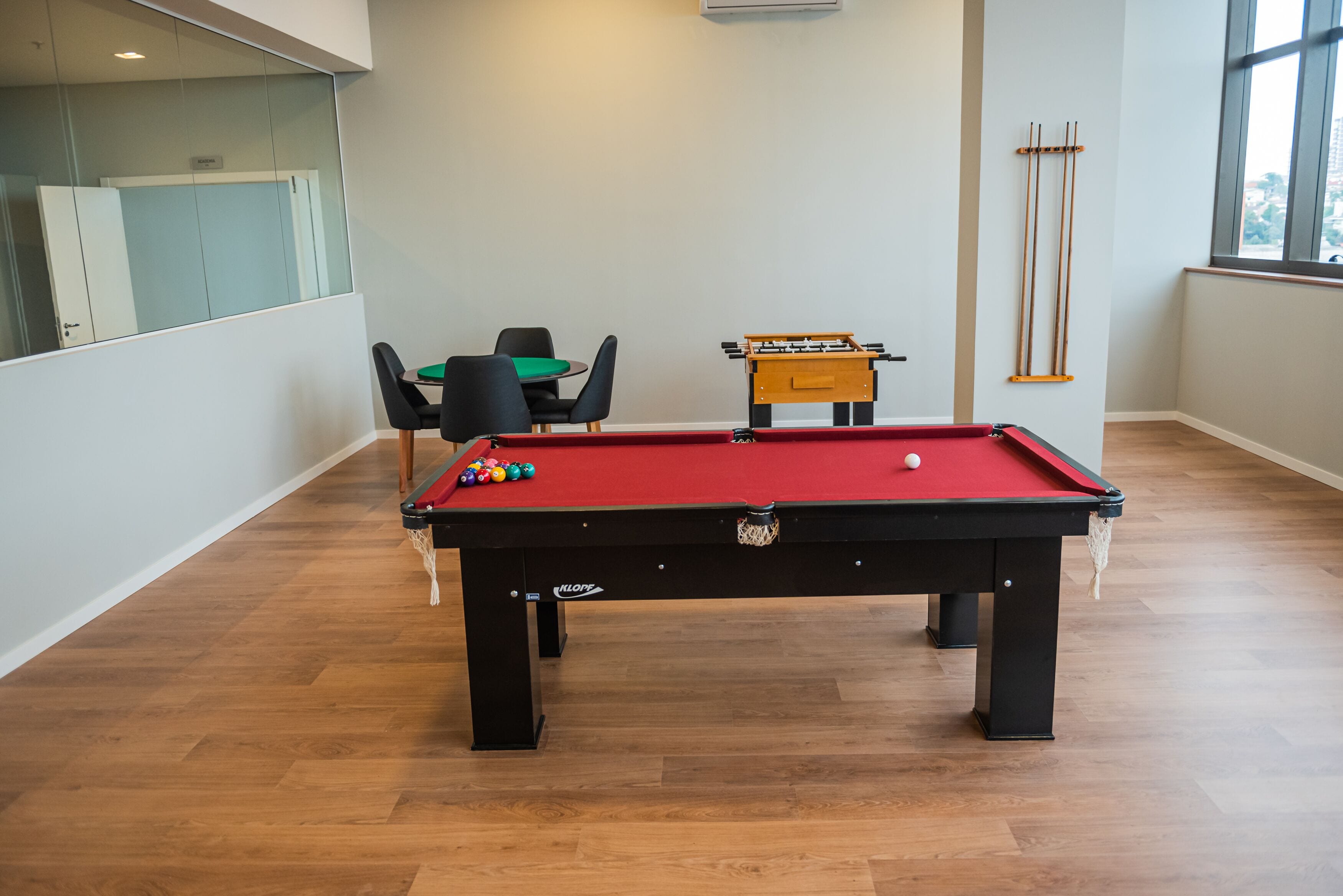 A pool table with balls, a foosball table, and a billiards rack in a room with wooden flooring and a glass partition.