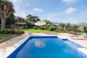 Ein Schwimmbecken mit Fliesenrand, umgeben von einem Garten mit Palmen und einer Pergola mit Sonnenschirm.