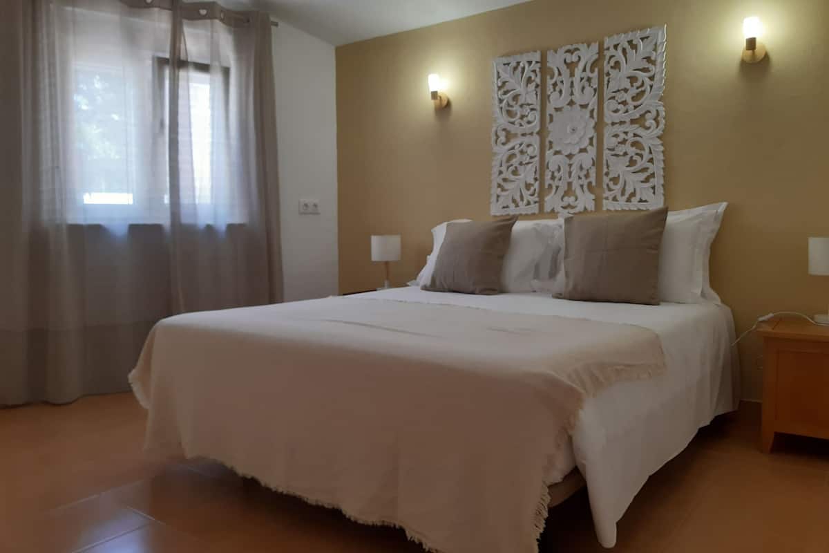 Une chambre à coucher avec un grand lit, deux lampes de chevet, un panneau décoratif au mur et une fenêtre ornée de rideaux légers.
