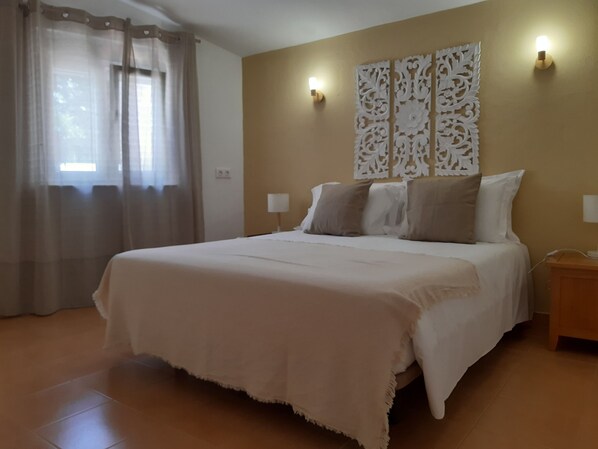 Un dormitorio con una cama grande, dos lámparas de noche, un panel decorativo en la pared y una ventana con cortinas translúcidas.