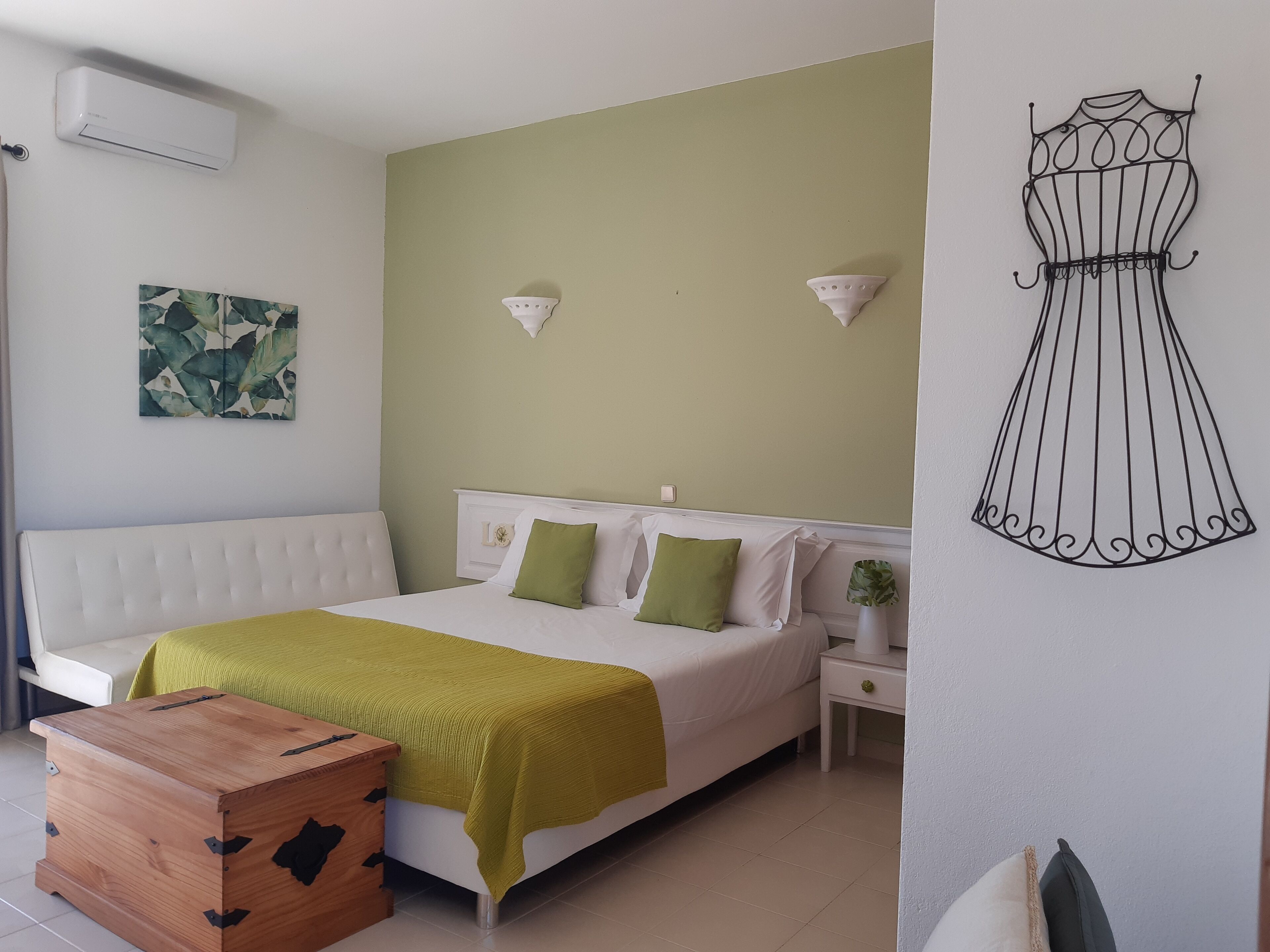 Quarto com cama, mesa de cabeceira, baú de madeira, luminária na parede e uma obra de arte na parede.