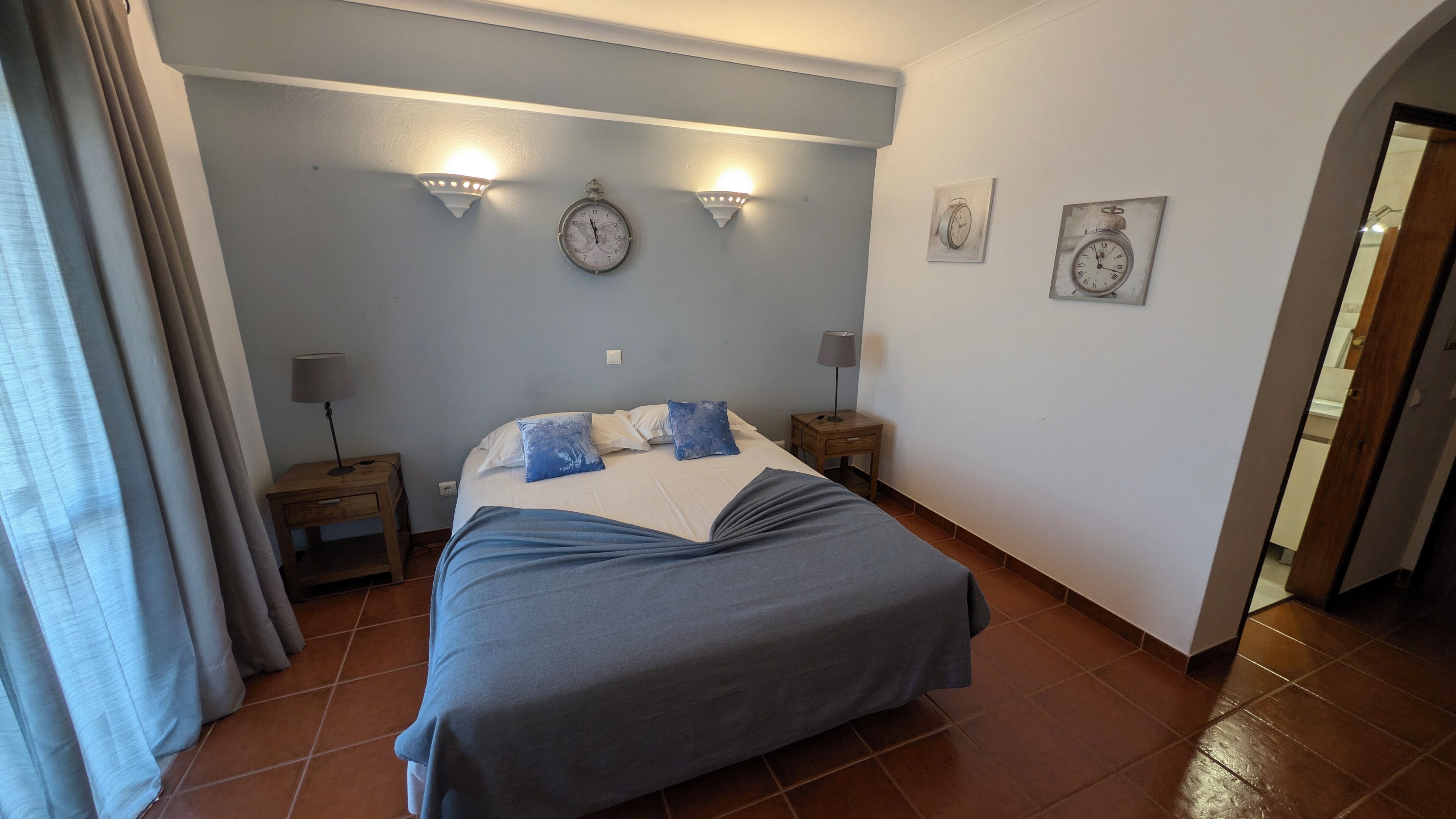 Une chambre à coucher comprenant un lit, deux tables de chevet, une horloge murale et deux lampes fixées au mur.