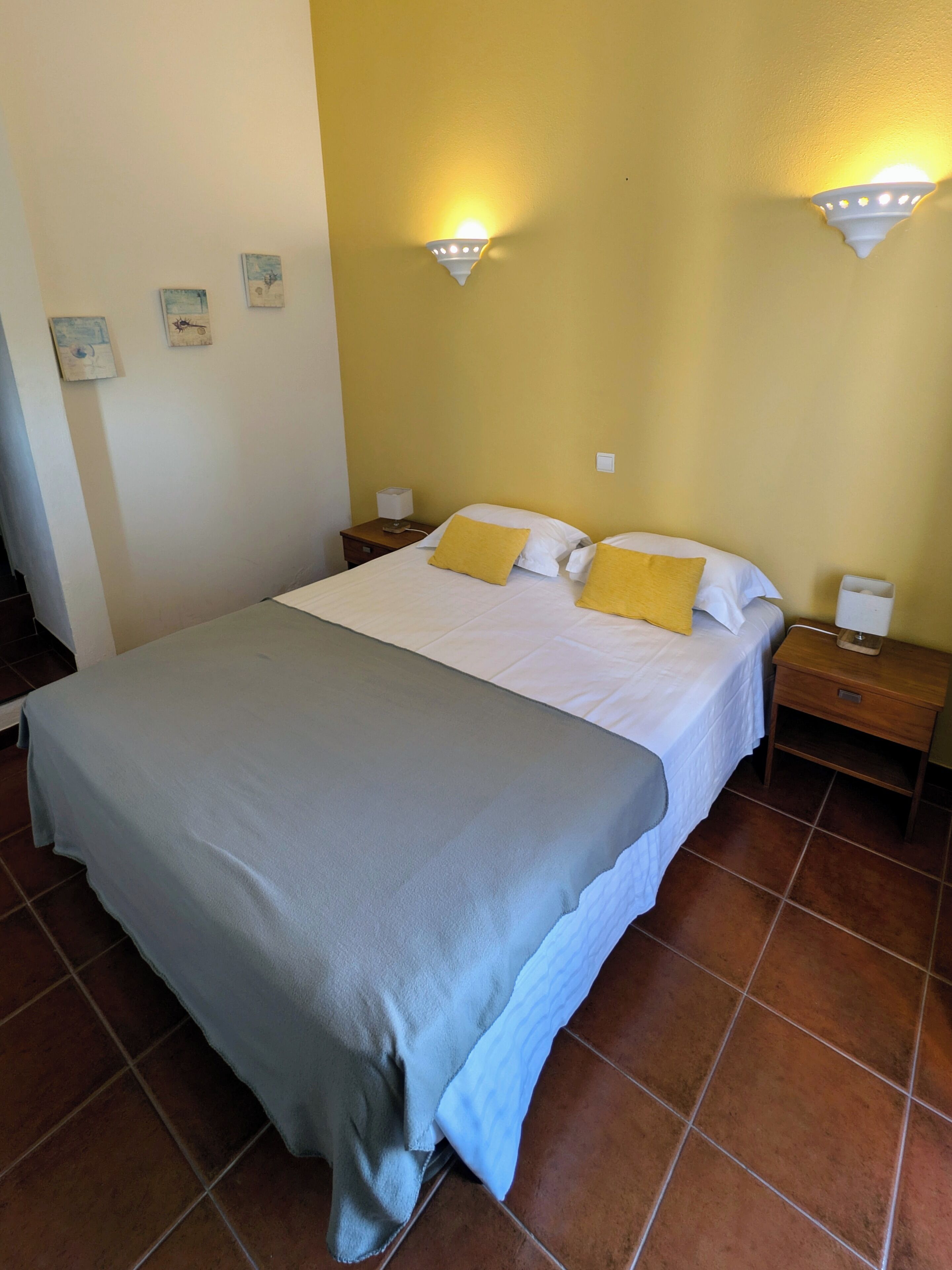 A neatly made bed with a grey blanket, two white pillows, and a yellow pillow. There are two bedside tables with lamps, a wall clock, and a small shelf with a telephone.
