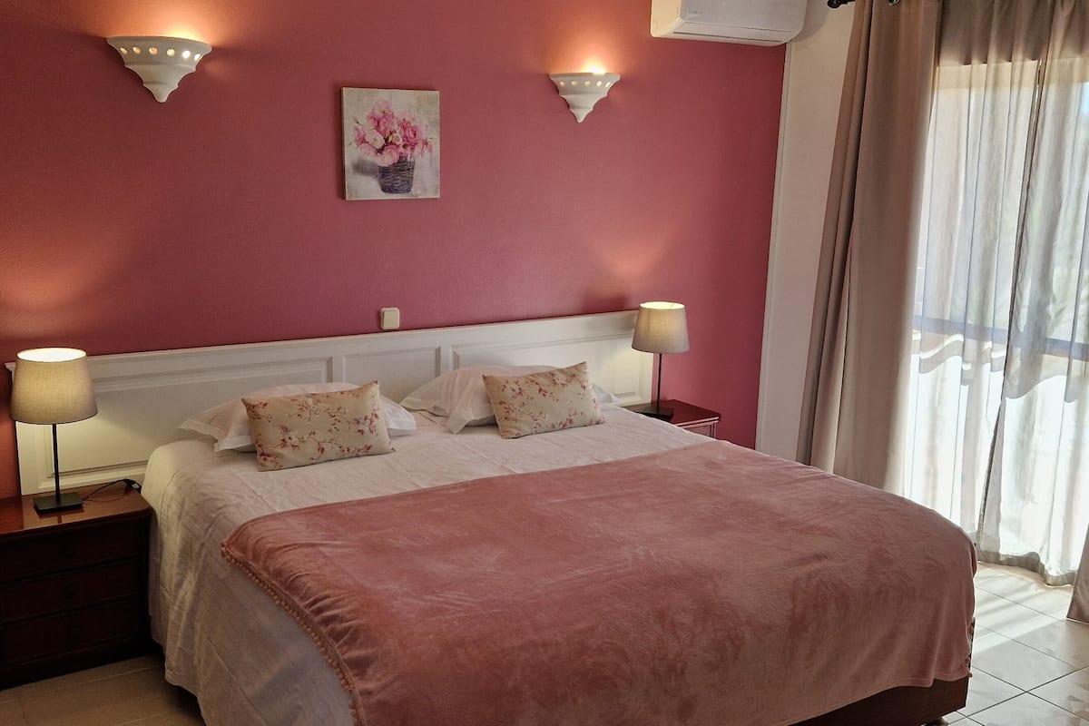 Un lit bien fait, recouvert d’une couverture rose, avec deux lampes de chevet et un motif floral décorant le mur.