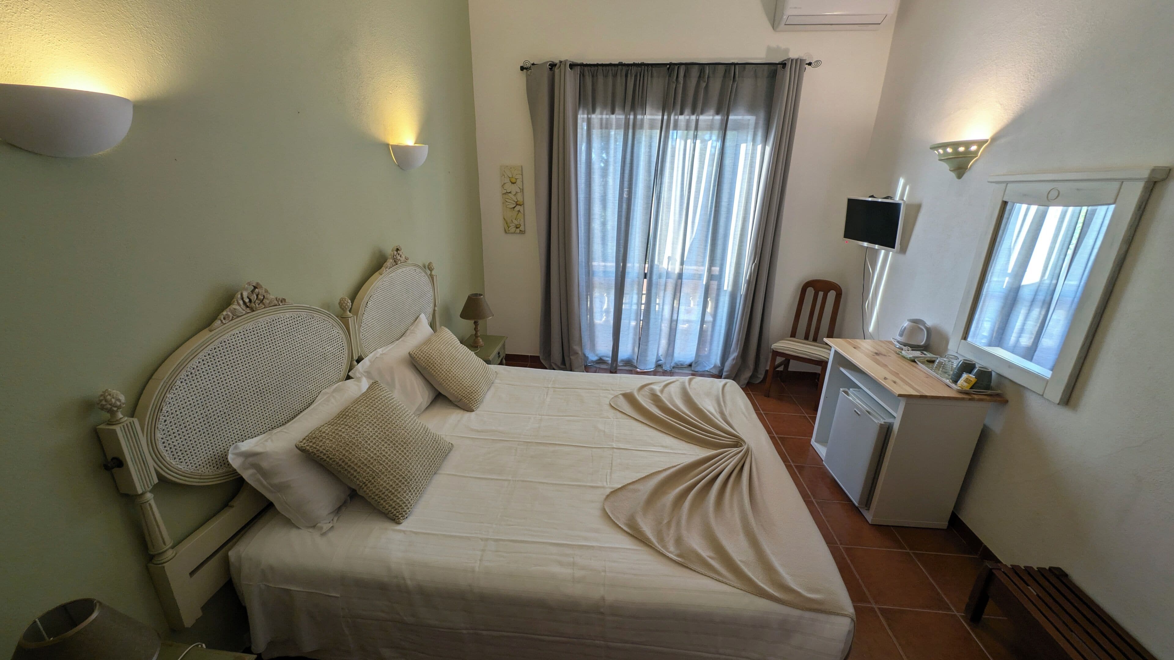 Uma cama bem arrumada com cabeceira, uma mesa de cabeceira com abajur, uma pequena escrivaninha com computador, uma cadeira e uma janela com cortinas.