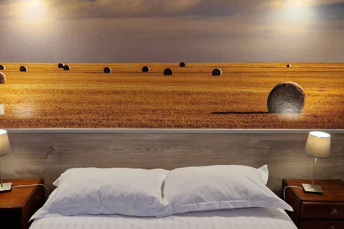 Une chambre à coucher avec un grand lit, deux tables de chevet avec des lampes et un tableau accroché au mur représentant un champ avec des bottes de foin.