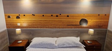 A bedroom with a large bed, two bedside tables with lamps, and a wall-mounted painting of a field with hay bales.