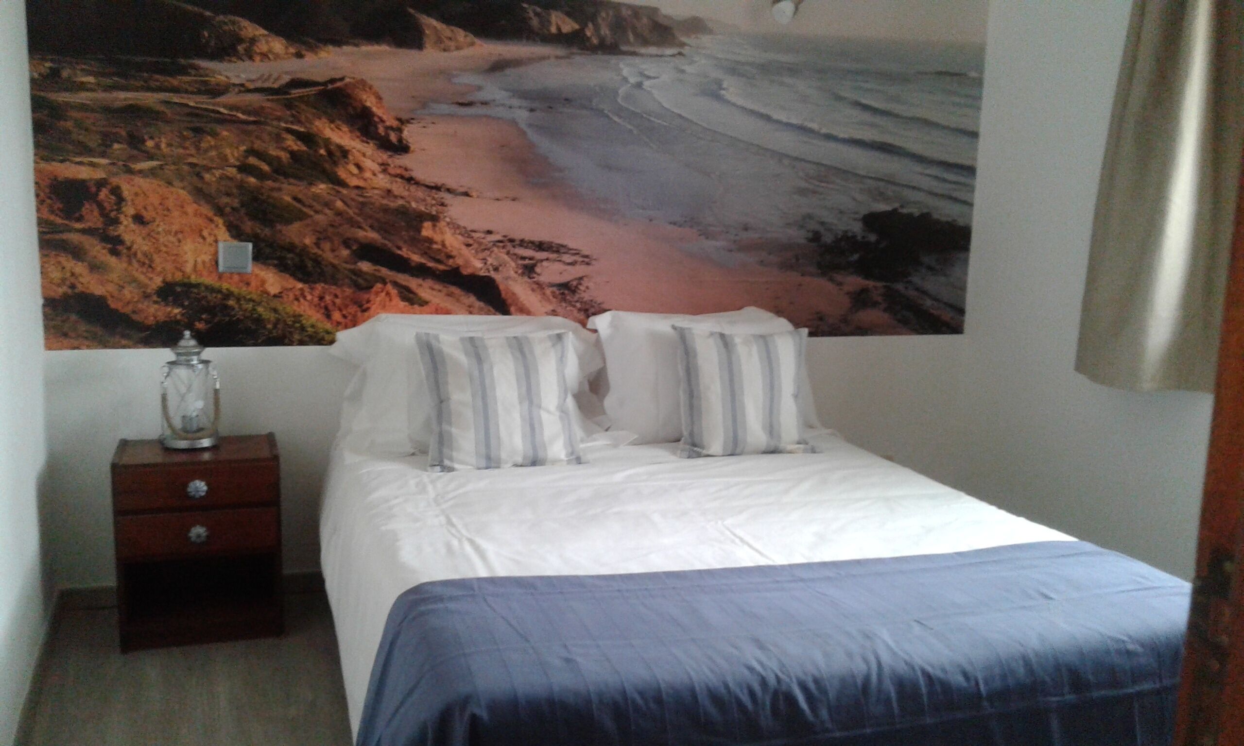 Ein Schlafzimmer mit Bett, Kissen, einem Nachttisch und Wandbild mit Strandmotiv.