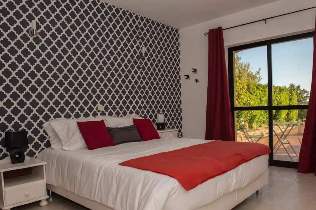 Une chambre avec un papier peint à motifs, un grand lit avec une literie rouge et blanche, une table de chevet avec une lampe, et une vue sur la verdure par une porte coulissante.