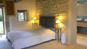 Une chambre avec un mur en pierre, un lit avec une literie blanche, deux lampes de chevet, un grand vase et une cheminée en pierre.