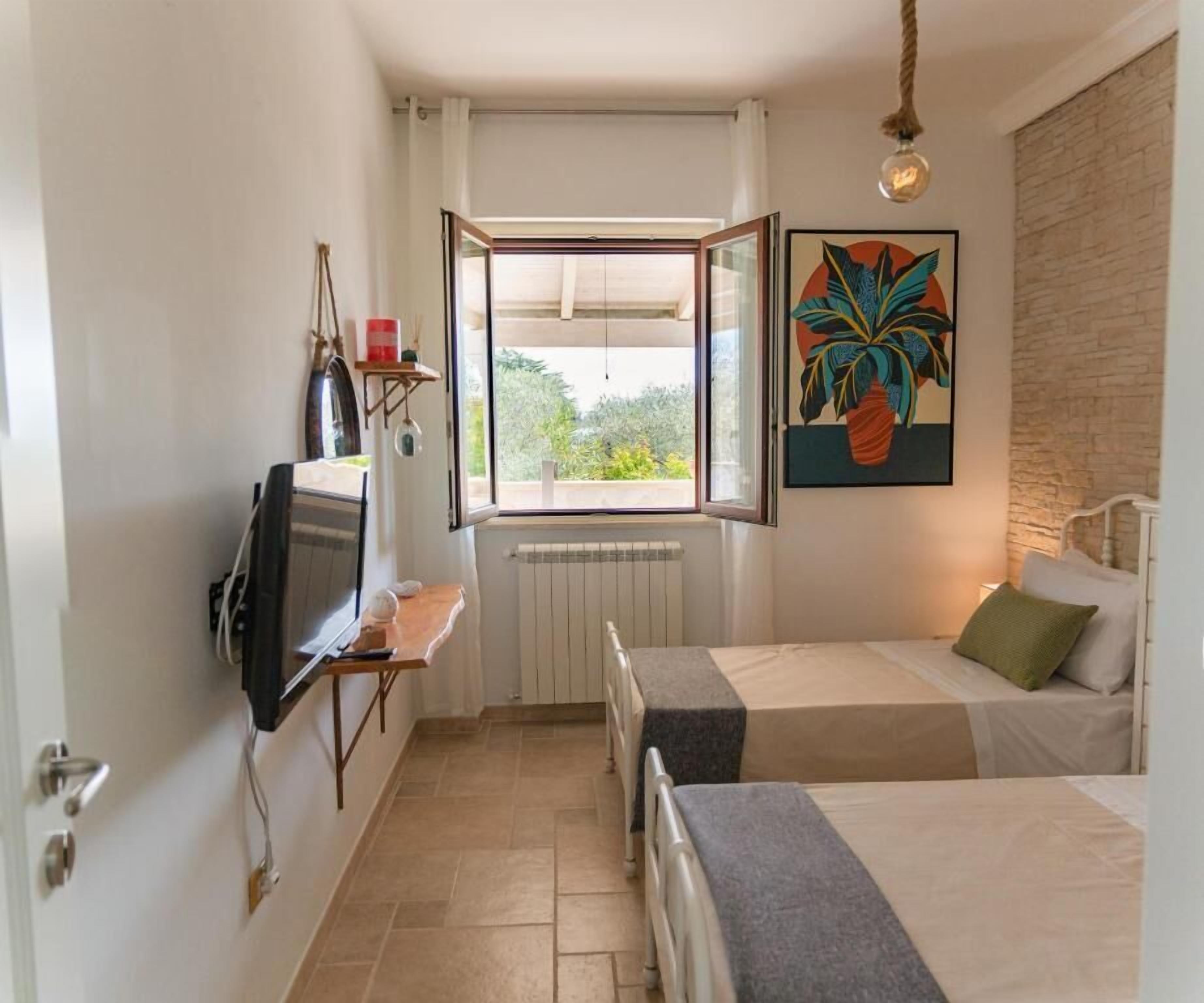 Une petite pièce bien éclairée, avec deux lits, une télévision, une fenêtre donnant sur un espace verdoyant et une plante décorative accrochée au mur.