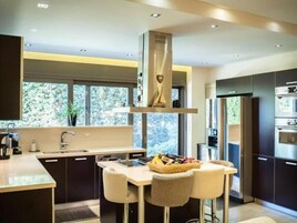 Una cocina moderna con armarios oscuros, una isla central con taburetes de bar, horno empotrado y un amplio ventanal con vistas a árboles.