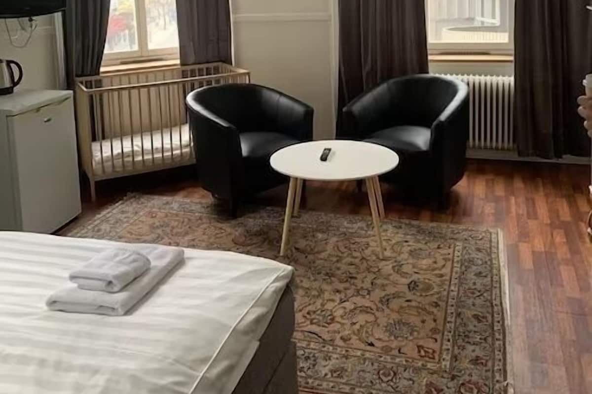 Habitación de hotel con cama, dos sillones negros, una mesita pequeña blanca, televisor, cuna y un ventanal grande con cortinas.