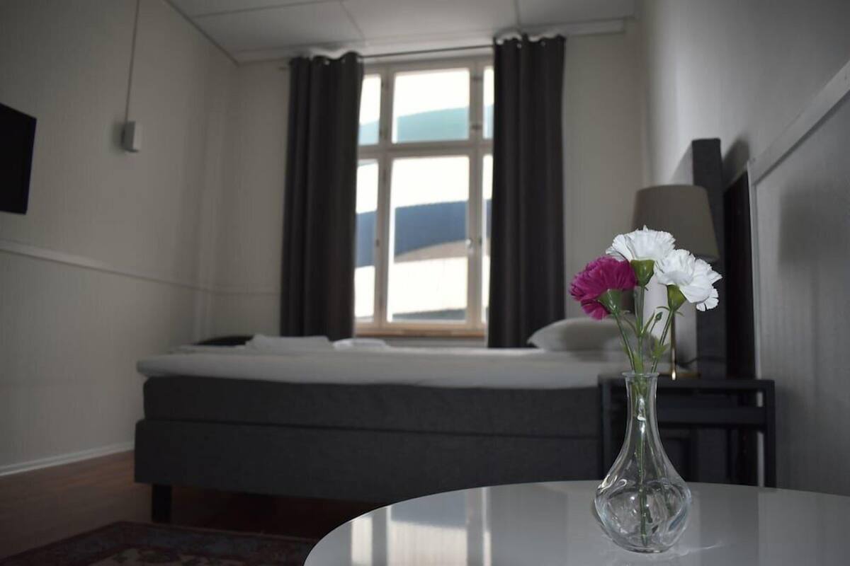Habitación de hotel con cama, un florero con flores, una ventana con cortinas y un televisor sobre un mueble.