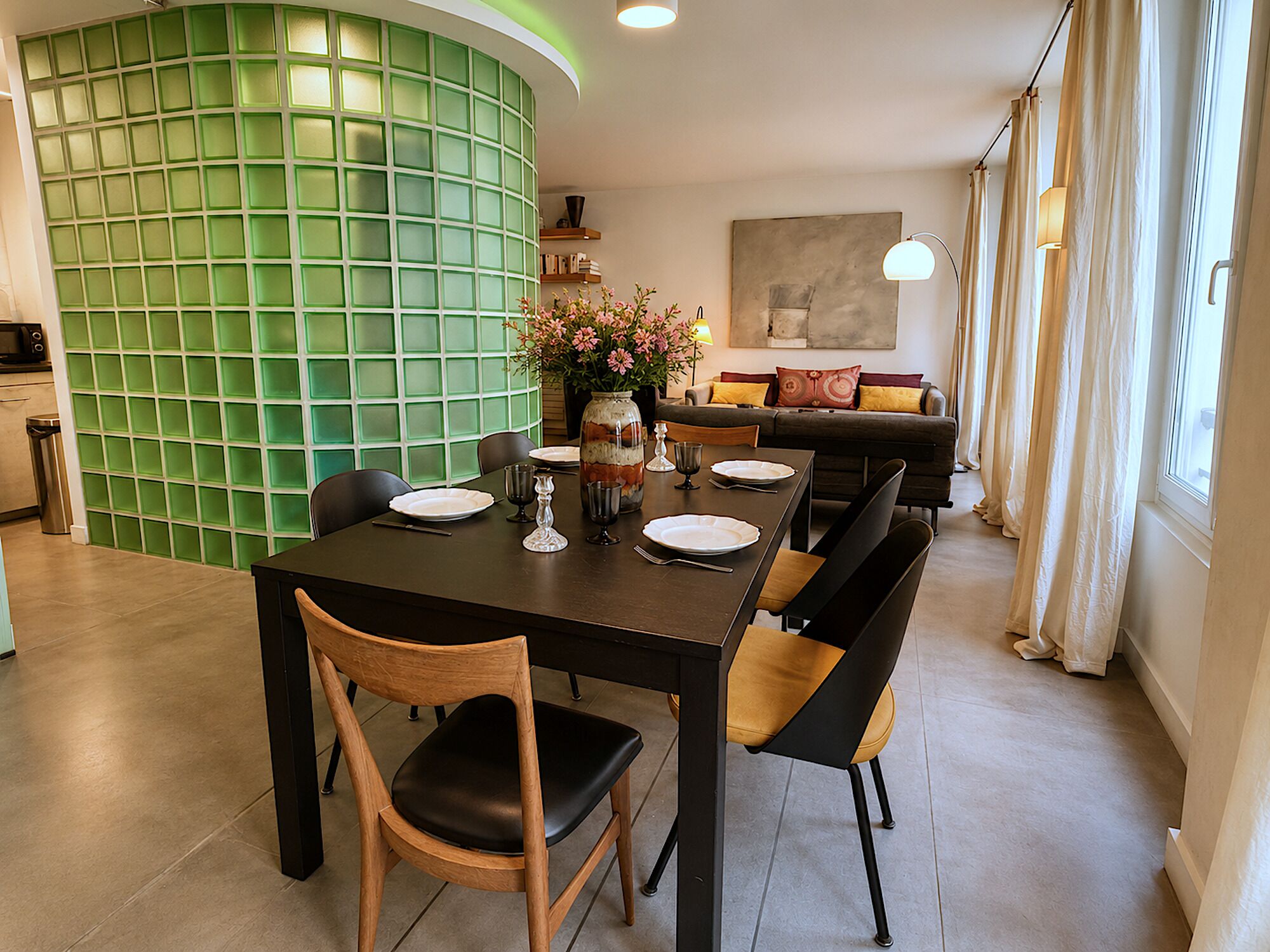 A dining area with a table set for four, surrounded by chairs, a vase with flowers, and a sofa in the background.