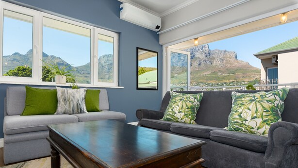 A living room with a sofa, armchairs, and a coffee table. Large windows offer a view of mountains and a building. - Vibrant Tamboerskloof Gem with Table Mountain View (Cape Town)