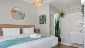 A modern hotel room with a large bed, a round mirror, a wall-mounted artwork, a bathroom with a shower, and a hanging plant. - Vibrant Tamboerskloof Gem with Table Mountain View (Cape Town)