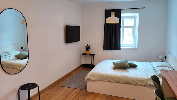 A modern bedroom with a bed, a desk, a chair, a TV, a window with curtains, a small table with a plant, and a mirror. - HIŠANAVOGALU (AP.3)Cozy studio in Lesce, near Bled (Radovljica)
