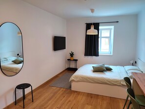 A modern bedroom with a bed, a desk, a chair, a TV, a window with curtains, a small table with a plant, and a mirror. - HIŠANAVOGALU (AP.3)Cozy studio in Lesce, near Bled (Radovljica)
