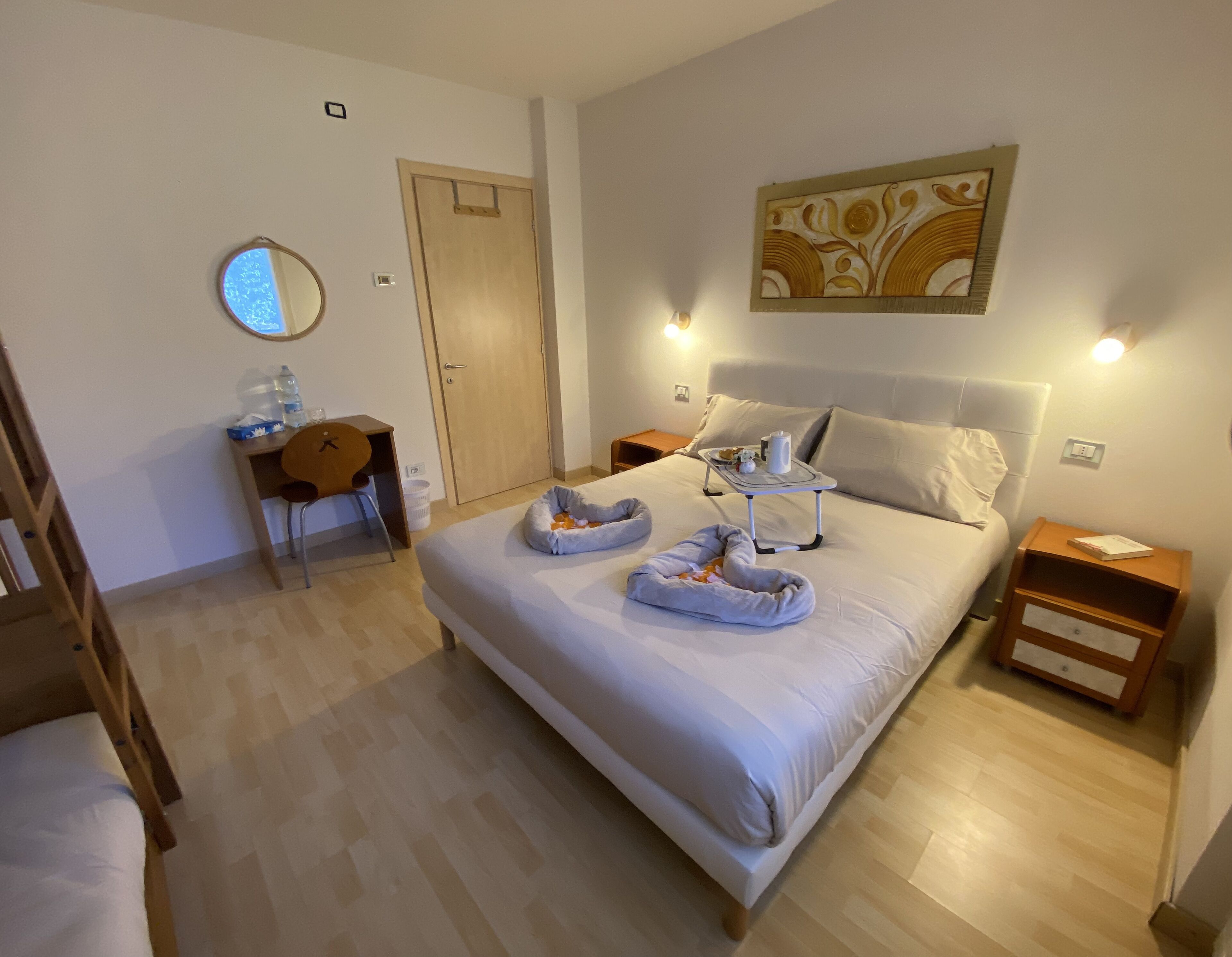 Habitación de hotel ordenada con una cama grande, mesitas de noche, un escritorio con silla, una lámpara de escritorio pequeña y un espejo montado en la pared.
