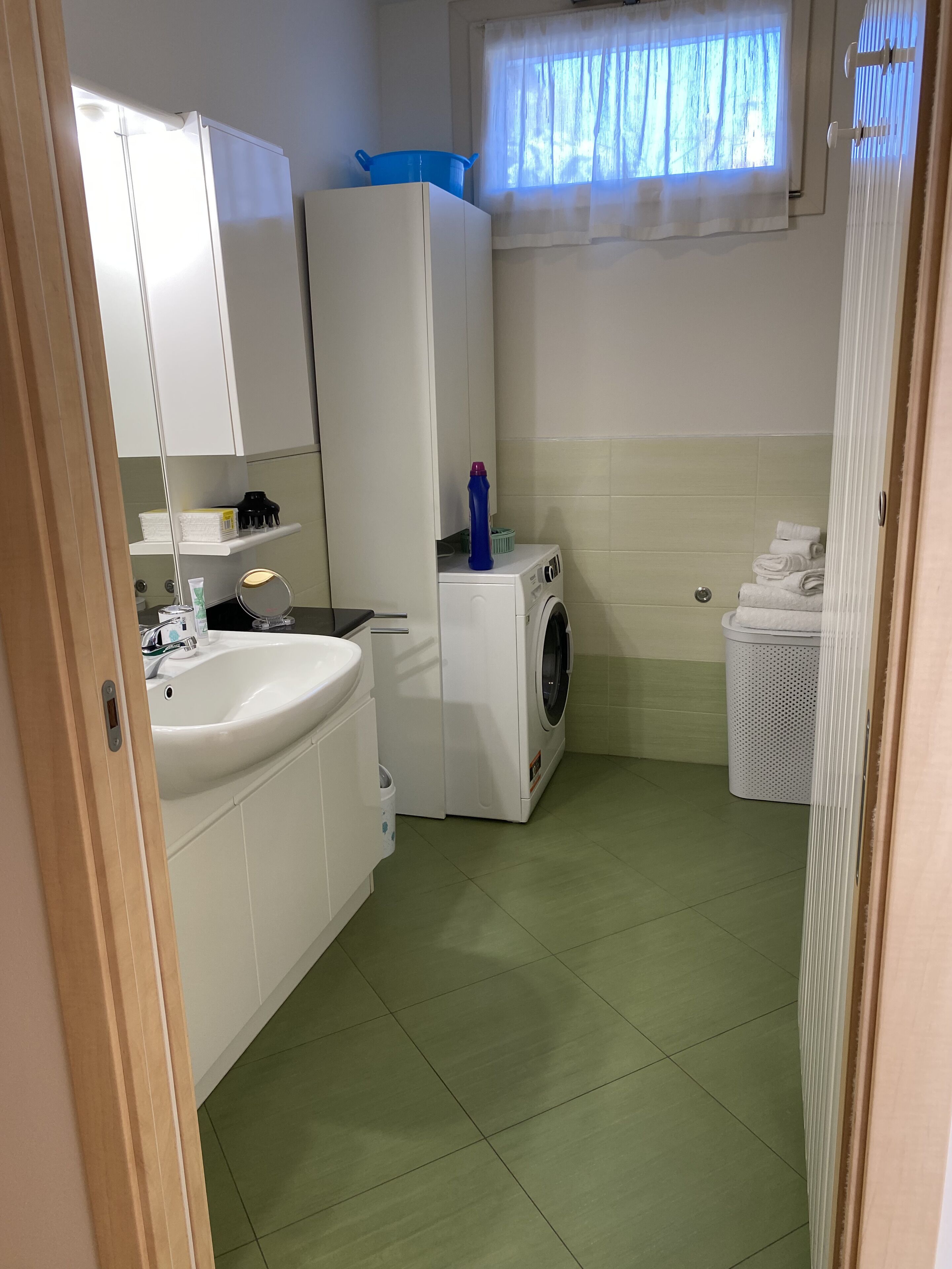 Ein kleines, gut eingerichtetes Badezimmer mit Waschbecken, Spiegel, weißen Schränken, einer Waschmaschine und einem Fenster mit Vorhängen.