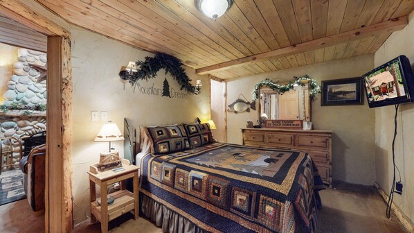 Quarto aconchegante com teto de madeira, cama com colcha estampada, cômoda, espelho e uma mesa pequena com abajur.