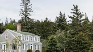 Une maison à deux étages, dotée d’une cheminée en pierre, entourée d’une forêt dense et bordée d’un rivage rocheux.